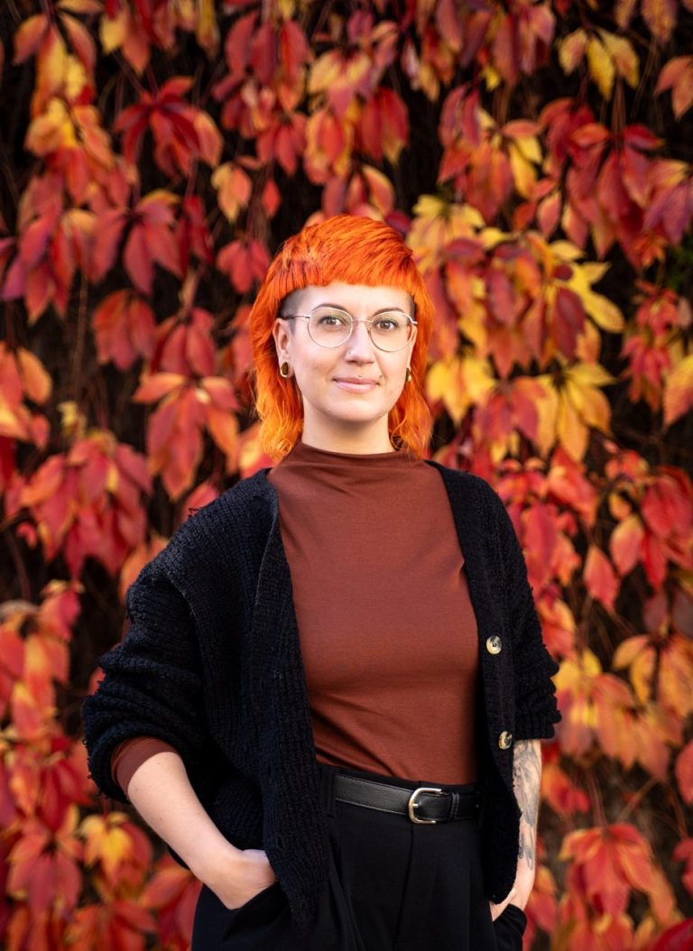 Jule Jule hat orangenes Haar und steht vor roten und gelben Weinblättern. Jule schaut freundlich in die Kamera.