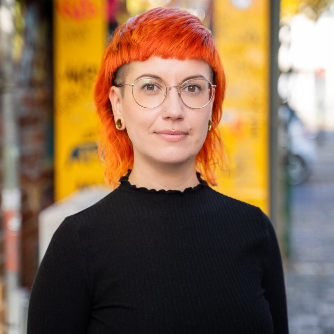 Jule im Hof Jule steht im Werk2 Hof vor einer alten gelben Telefonzelle. Jule schaut freundlich in die Kamera. Jule trägt eine Brille.