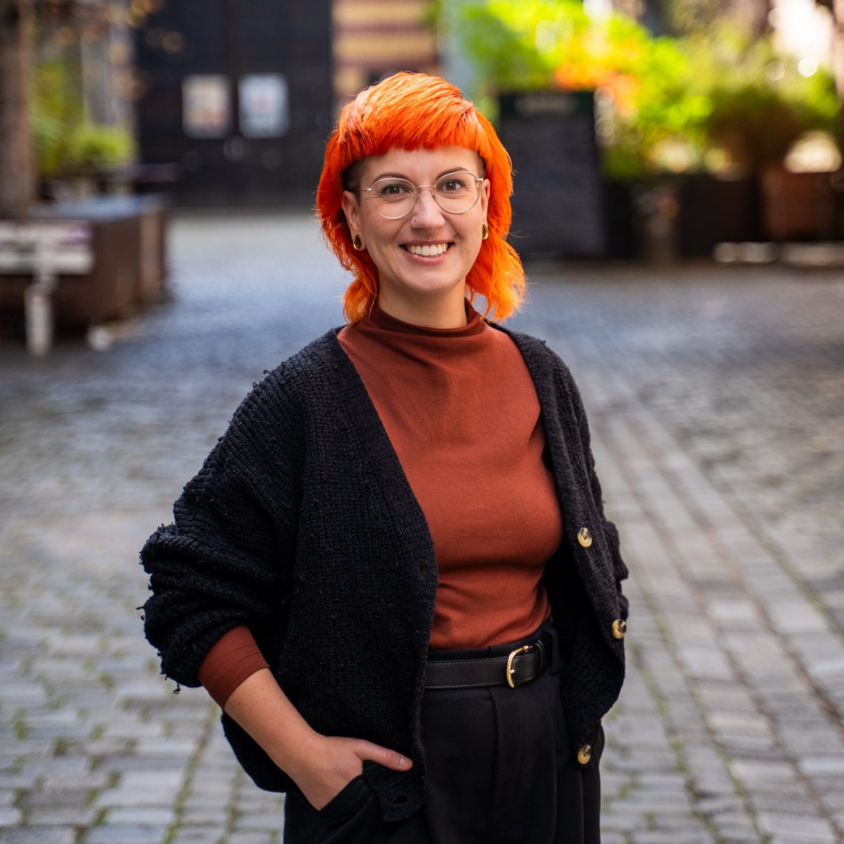 Jule hat orangefarbenes Haar, trägt ein braunes Oberteil und einen schwarzen Cardigan und lächelt.