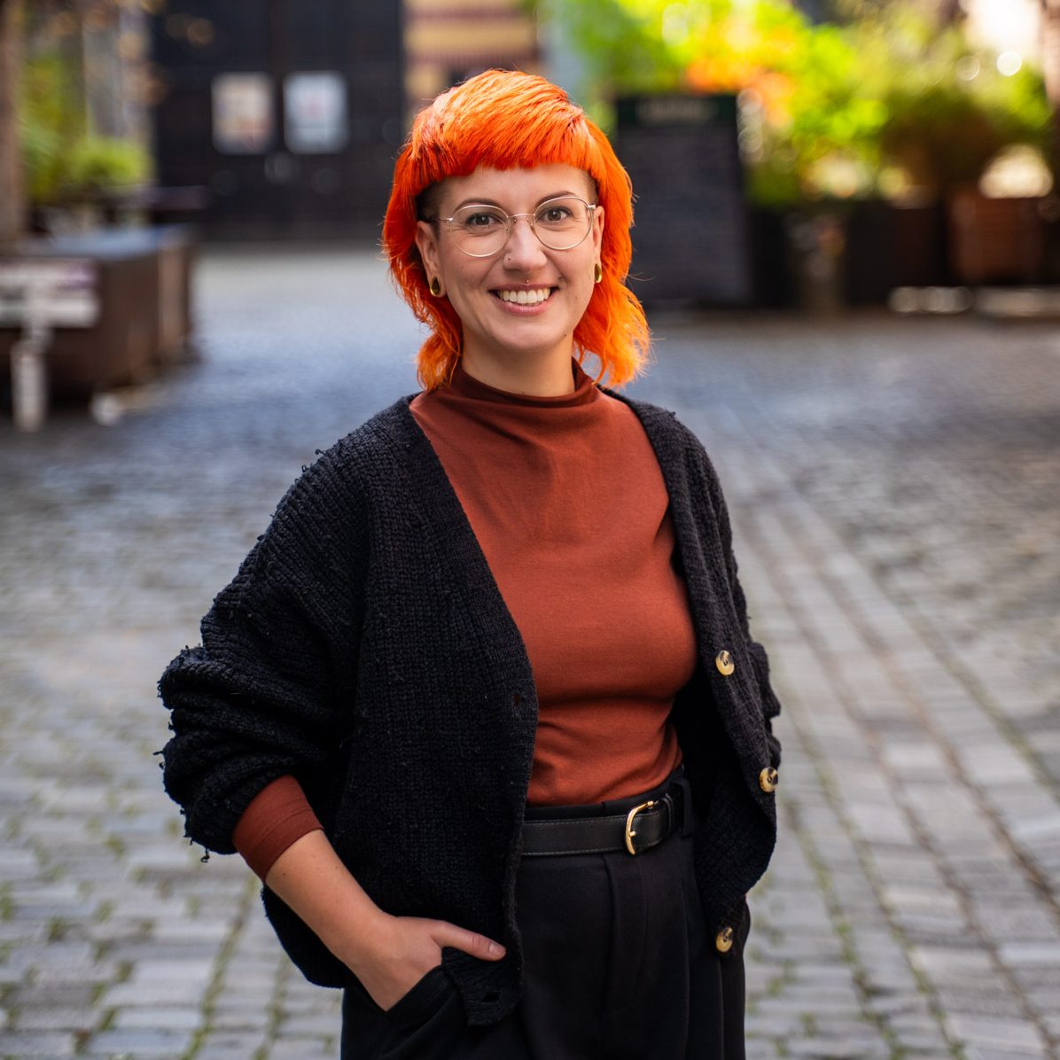 Jule steht im Hof vom Werk2 und schaut lachend in die Kamera. Jule hat orangene Haare und trägt ein braunes Oberteil mit schwarzem Cardigan
