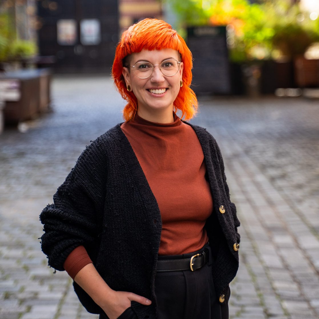 Frau mit orangefarbenem Haar und Brille trägt einen schwarzen Cardigan und steht lächelnd im Freien.