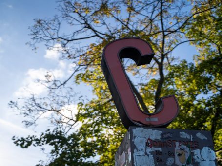 Roter, großer Buchstabe "C" vor blauem Himmel und grünen Ästen. Das ist der Hofeingang zur Praxis.